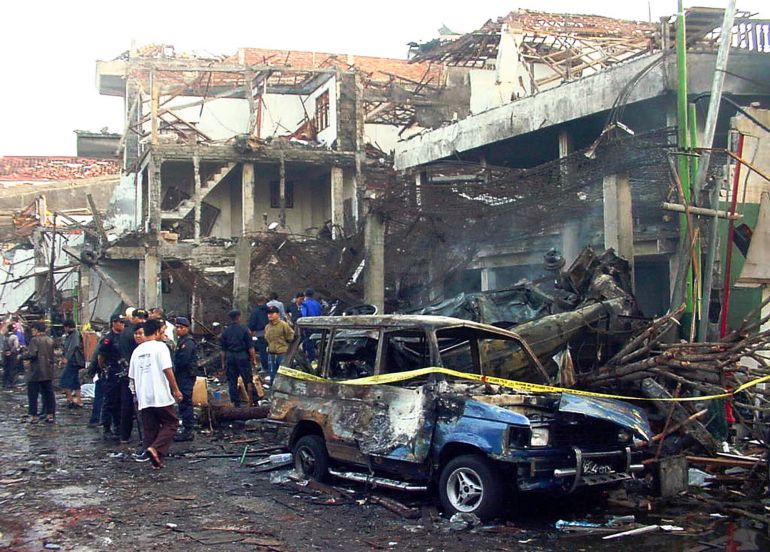 The scene of the Bali bombings in 2002.. Police officers are inspecting the ruins of the devastated buildings. Others are watching. A wrecked car is in front. It has yellow police tape around it.