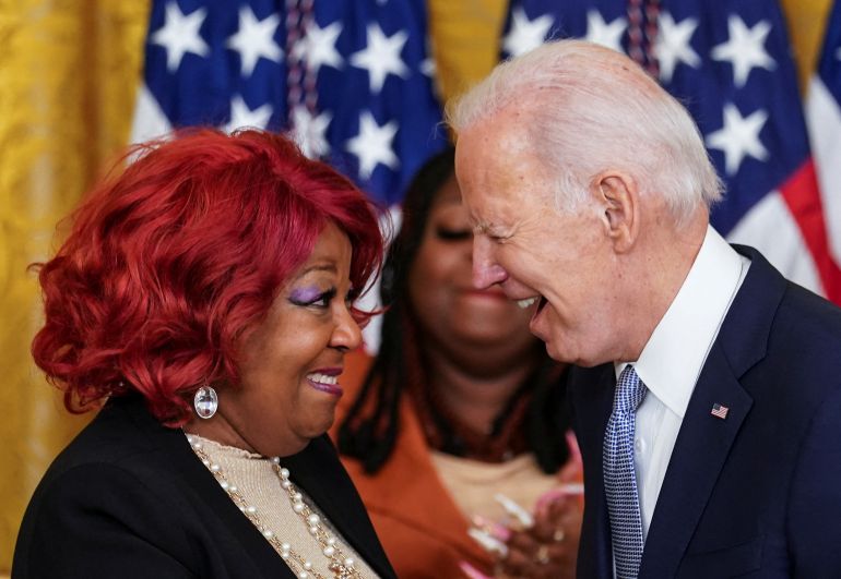 President Joe Biden leans over to speak to Ruby Freeman, as her daughter applauds behind them. In the background are two US flags.