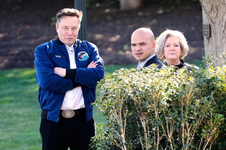 Elon Musk crosses his arms, next to Walt Nauta and Susie Wiles, standing on the White House lawn.