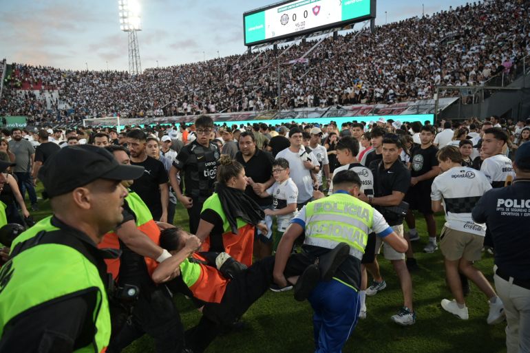 A police officer carries a fan of Cerro Porteño as they walk on the pitch after clashes with police officers during the Paraguayan tournament football match between Olimpia and Cerro Porteño at the Defensores del Chaco Stadium in Asunción on April 19, 2026. Several people were injured and at least a hundred were arrested following incidents that occurred during the Paraguayan football derby between Olimpia and Cerro Porteno, which was suspended in the first half in Asuncion on April 19, 2026. (Photo by Daniel DUARTE / AFP)